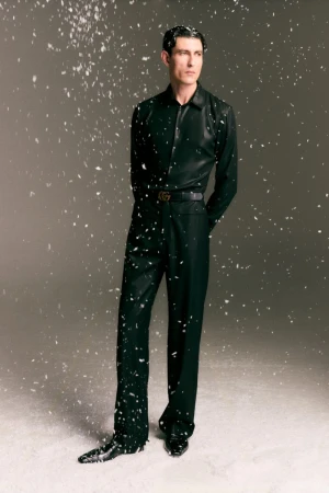 Model standing in dark trousers and matching shirt against a grey gradient backdrop with falling snow particles