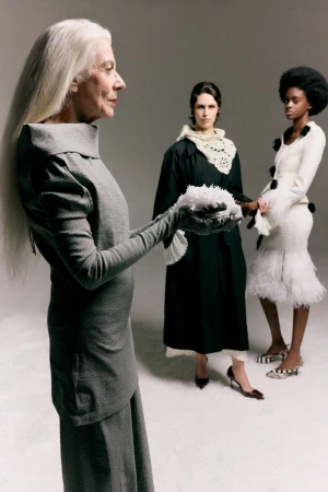 Three models in styled garments standing in a studio with an elderly woman holding a feathery cluster in the foreground profile.