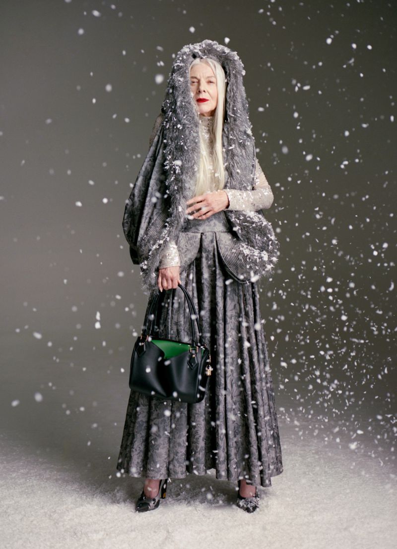 Model in textured grey hooded coat holding a black handbag amidst falling snow against a plain grey background