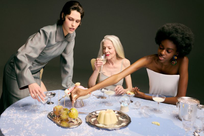 Three female models gathered around a light blue table laden with desserts, drinks, and silver platters against a dark grey studio background.