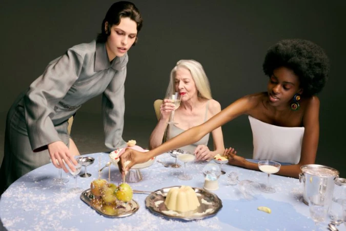 Three female models gathered around a light blue table laden with desserts, drinks, and silver platters against a dark grey studio background.
