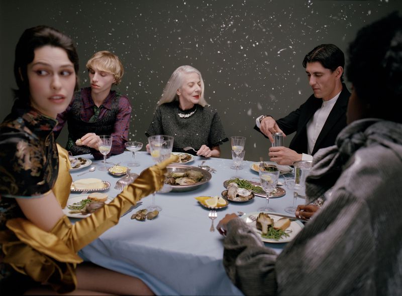Group of people seated around a dining table with a light blue tablecloth against a dark grey background with falling snow effects