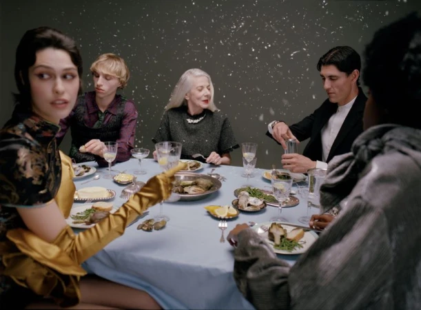 Group of people seated around a dining table with a light blue tablecloth against a dark grey background with falling snow effects