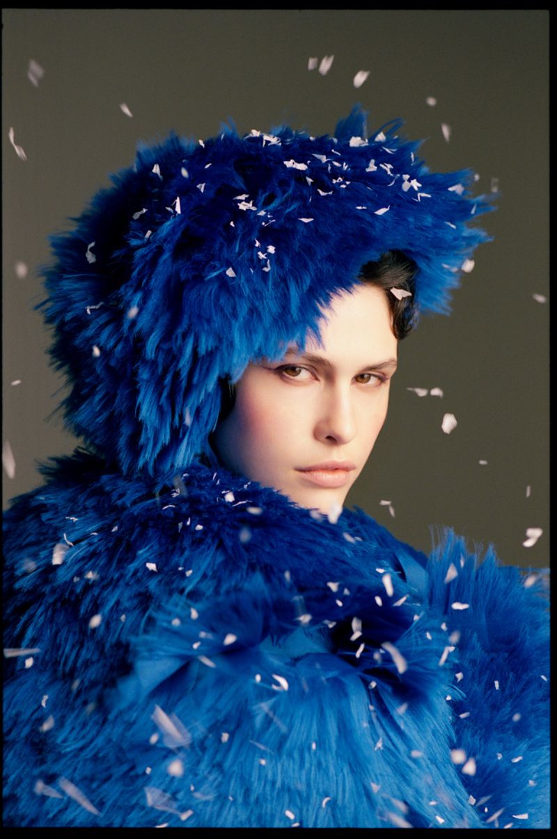 Model wearing a textured blue hooded garment with scattered white confetti flakes falling around them