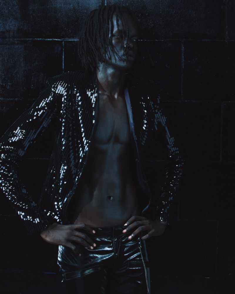 Man wearing a dark sequined jacket and dark trousers standing against a dark tiled background