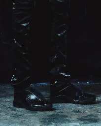 Close-up of shiny black sneakers and glossy black trousers against a dark textured ground