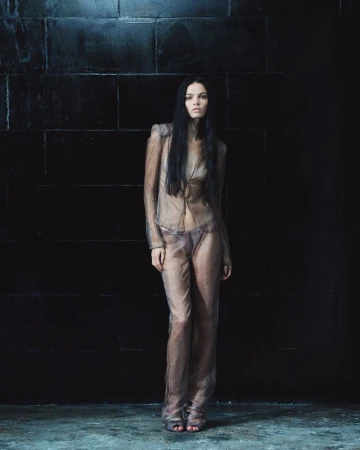 Model with long dark hair standing against a dark tiled wall in a sheer taupe outfit for Mowalola