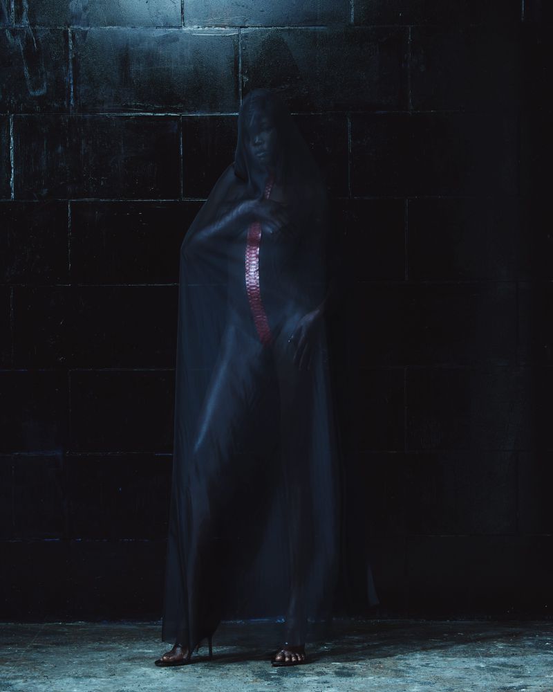 Model draped in sheer dark fabric standing before a dark brick wall for Mowalola