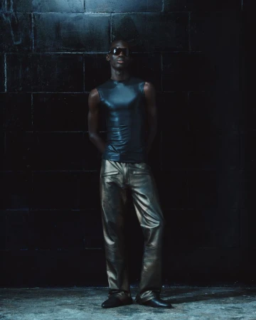 Model wearing dark sunglasses, sleeveless top, and metallic trousers standing against a dark tiled wall.