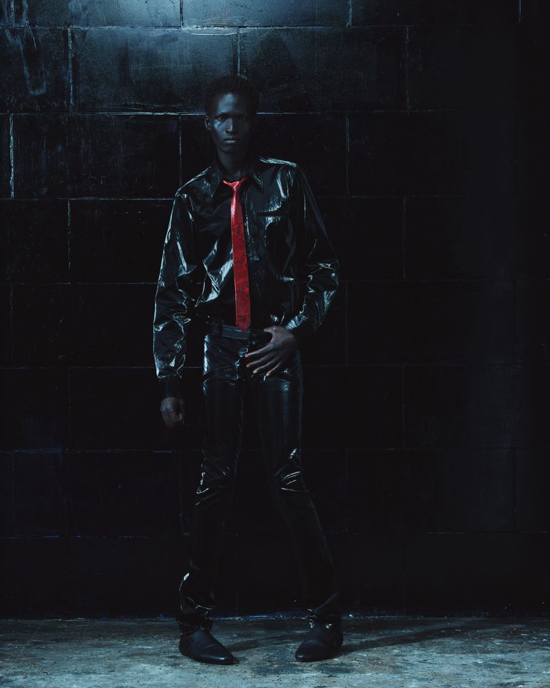 Model standing against a dark brick wall wearing a glossy black shirt, matching trousers, and a bright red tie.