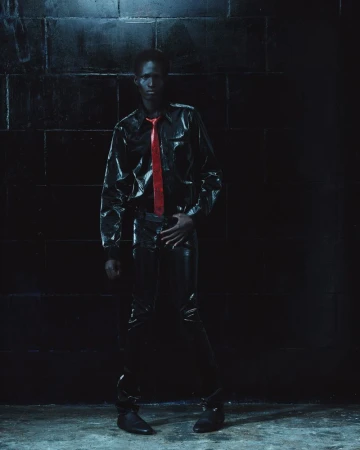 Model standing against a dark brick wall wearing a glossy black shirt, matching trousers, and a bright red tie.