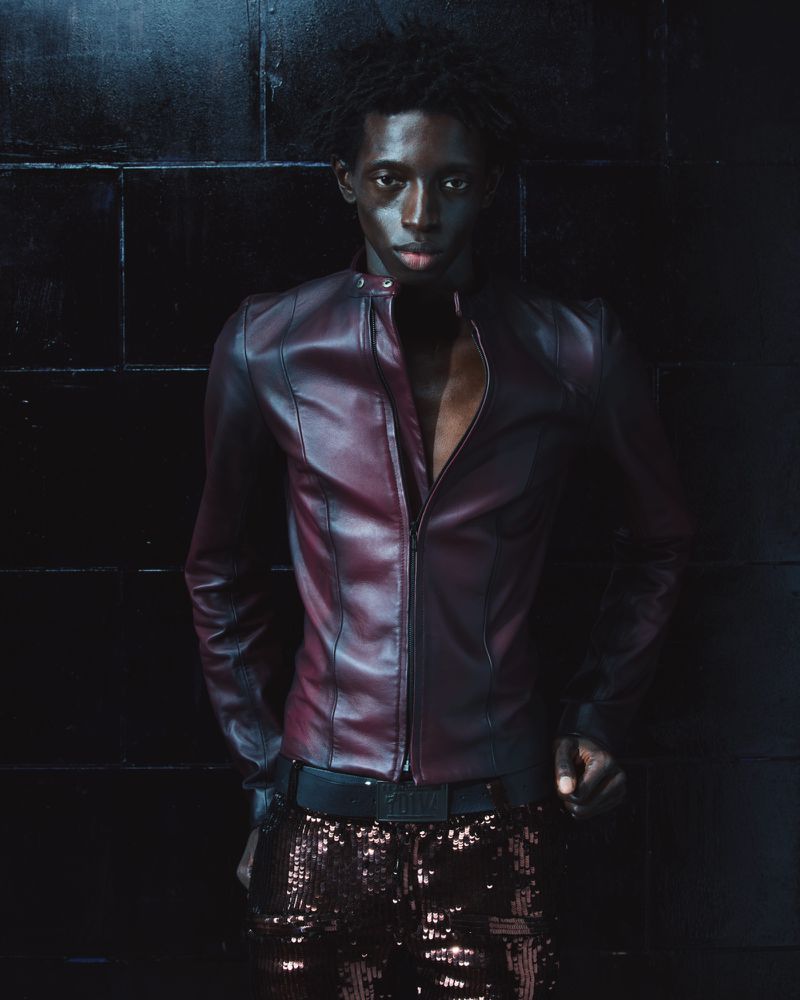 Black man wearing a burgundy leather jacket and dark sequin pants standing against a dark tiled wall.