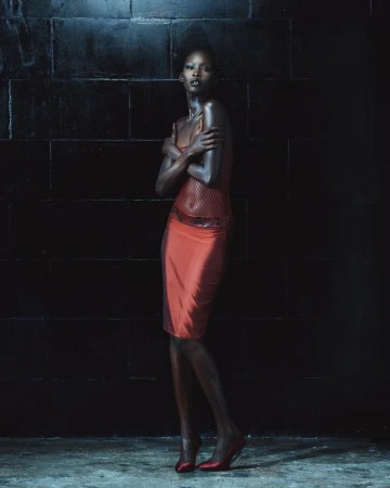 Model wearing a red slip skirt and matching heels standing against a dark tiled wall in a moody setting.