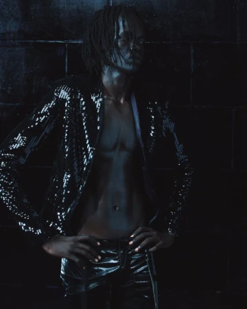 Man wearing a dark sequined jacket and dark trousers standing against a dark tiled background