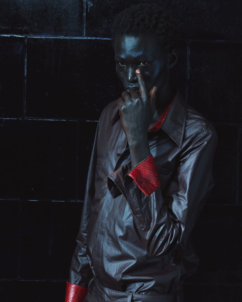 Portrait of a black man wearing a dark shirt with red cuffs posing against a black tiled wall