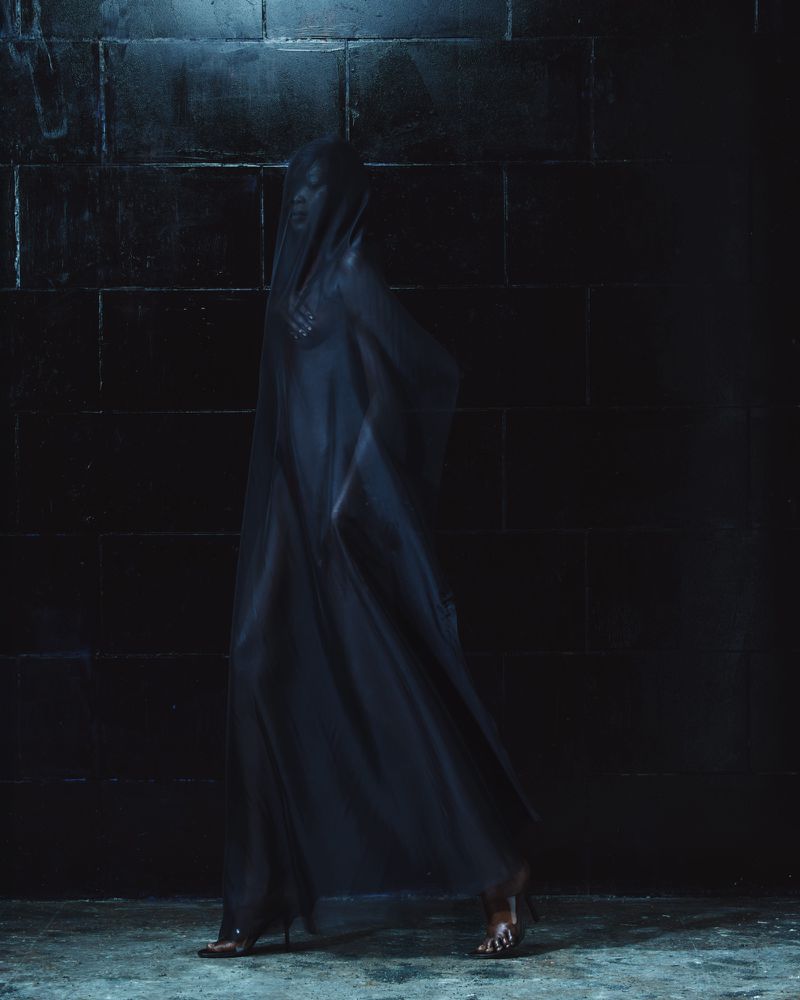 Model draped in a sheer black veil standing against a dark tiled wall for Mowalola