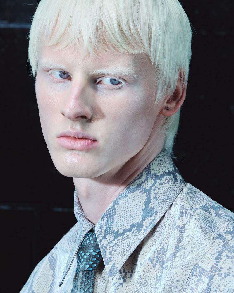 Close-up portrait of a young man with platinum blonde hair wearing a snakeskin print shirt and patterned tie for Mowalola.