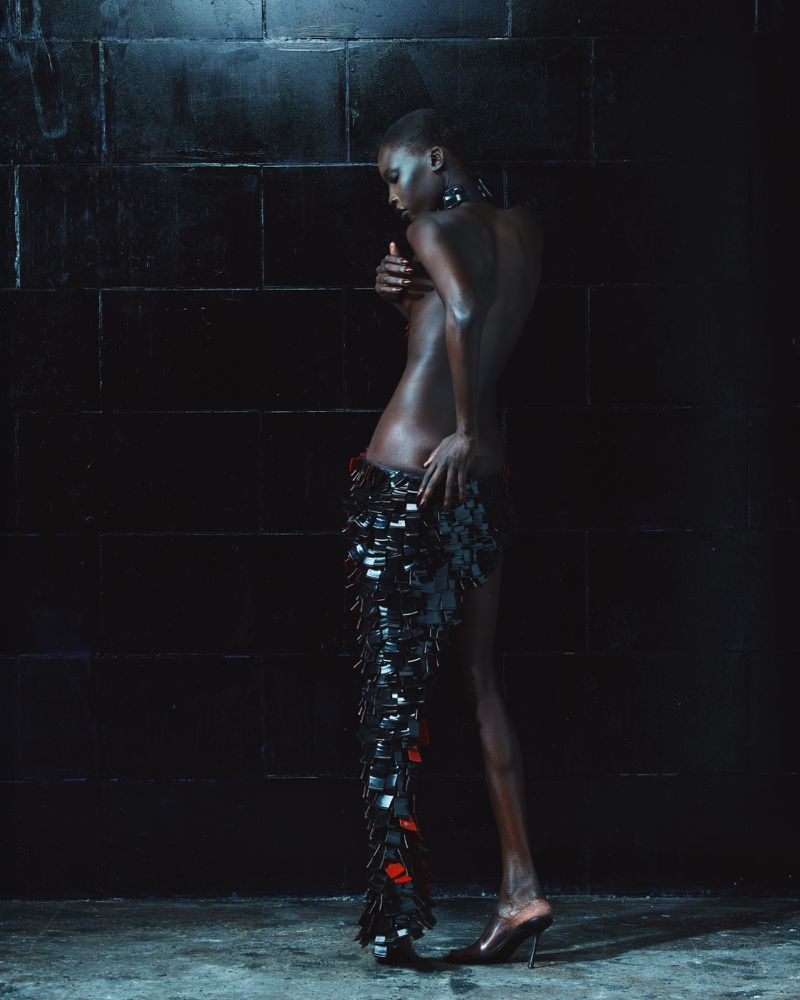 Model wearing a textured dark skirt and heels standing against a dark tiled wall in dramatic low-key lighting for Mowalola