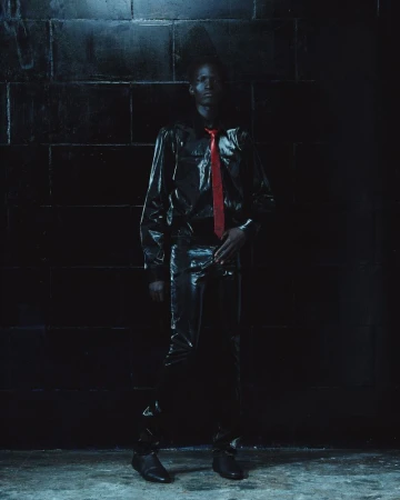 Model wearing dark glossy shirt and trousers with a red tie against a dark tiled background