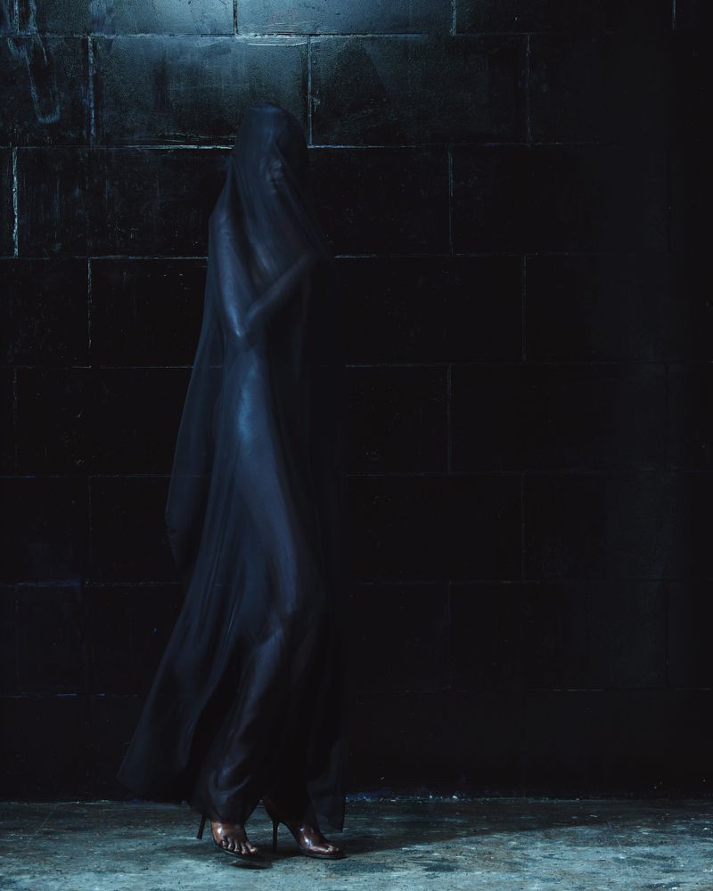 A veiled figure in a dark flowing gown standing against a dark brick wall in moody lighting.