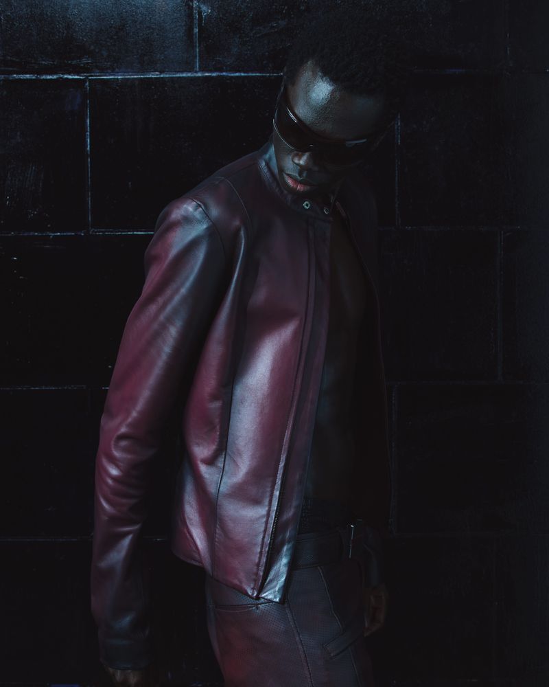 Model wearing a dark leather jacket and sunglasses standing against a dark tiled background for Mowalola lookbook