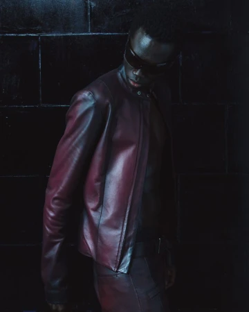 Model wearing a dark leather jacket and sunglasses standing against a dark tiled background for Mowalola lookbook