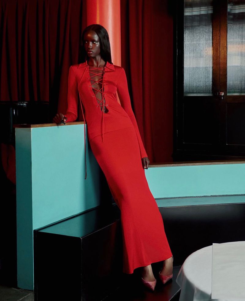 Model in a long red lace-up dress leaning against a turquoise partition in a dimly lit restaurant interior