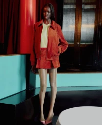 Model Mayor Dutie wearing a rust-colored jacket and skirt standing inside a warmly lit interior space with red drapes and a dark door.