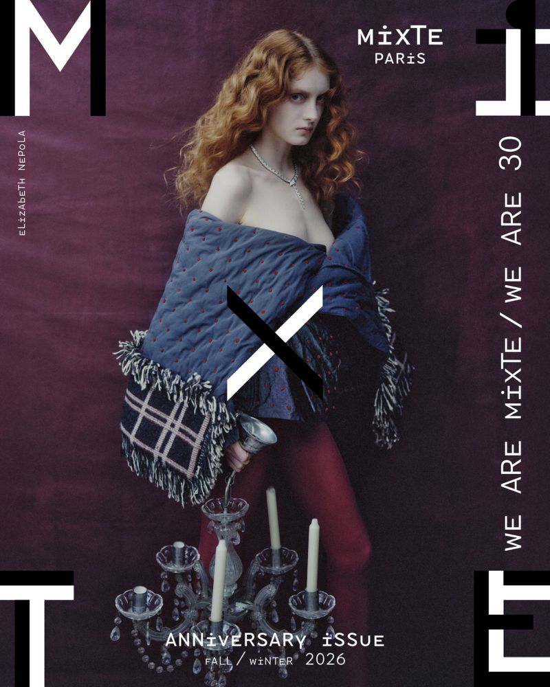 Model with long red curly hair wearing a blue quilted wrap and holding an ornate glass candelabra against a dark backdrop.