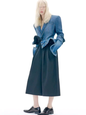 Model with long blonde hair wearing a structured denim jacket and dark trousers against a bright white background.