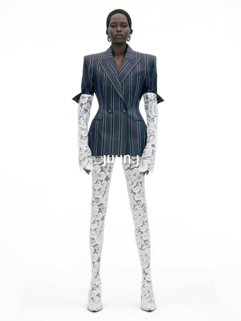 Model standing against a white seamless background wearing a pinstriped blazer and white lace sleeves