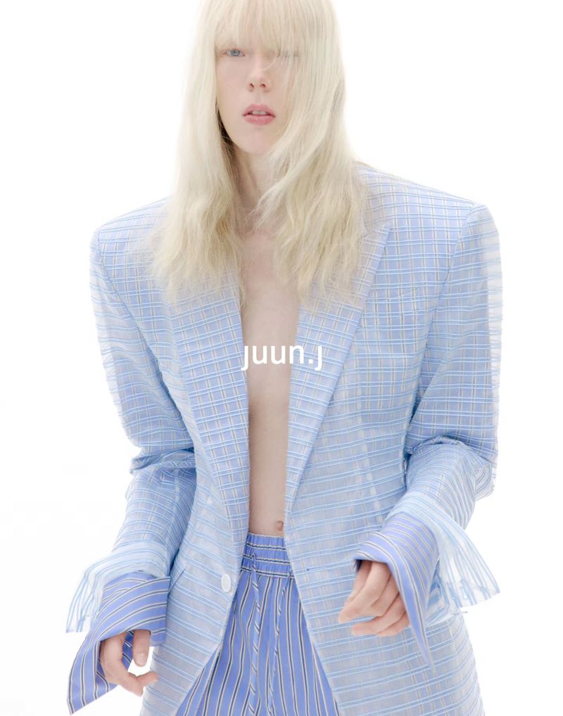 Model with long blonde hair wearing a light blue plaid blazer and striped trousers against a white background.