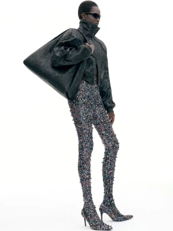 Model wearing dark sunglasses, black leather jacket, and embellished leggings carrying a large black shoulder bag against a white background
