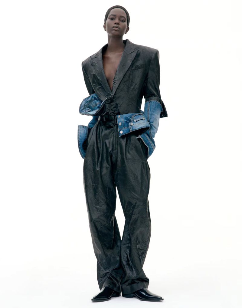 Model standing against a white backdrop wearing a dark structured suit with denim sleeve details