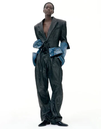 Model standing against a white backdrop wearing a dark structured suit with denim sleeve details