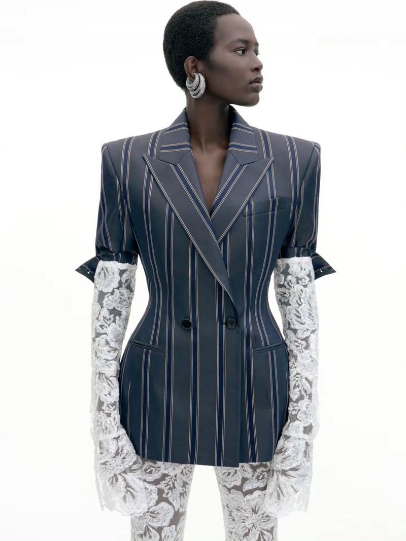 Model with short hair wearing a pinstripe double-breasted blazer and white lace gloves against a white background