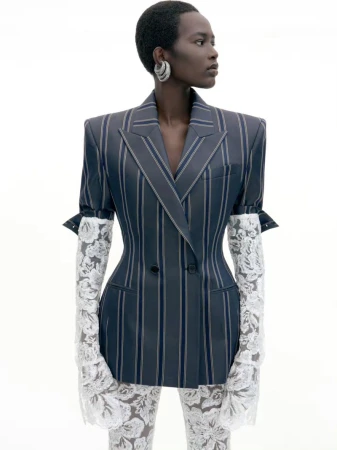 Model with short hair wearing a pinstripe double-breasted blazer and white lace gloves against a white background
