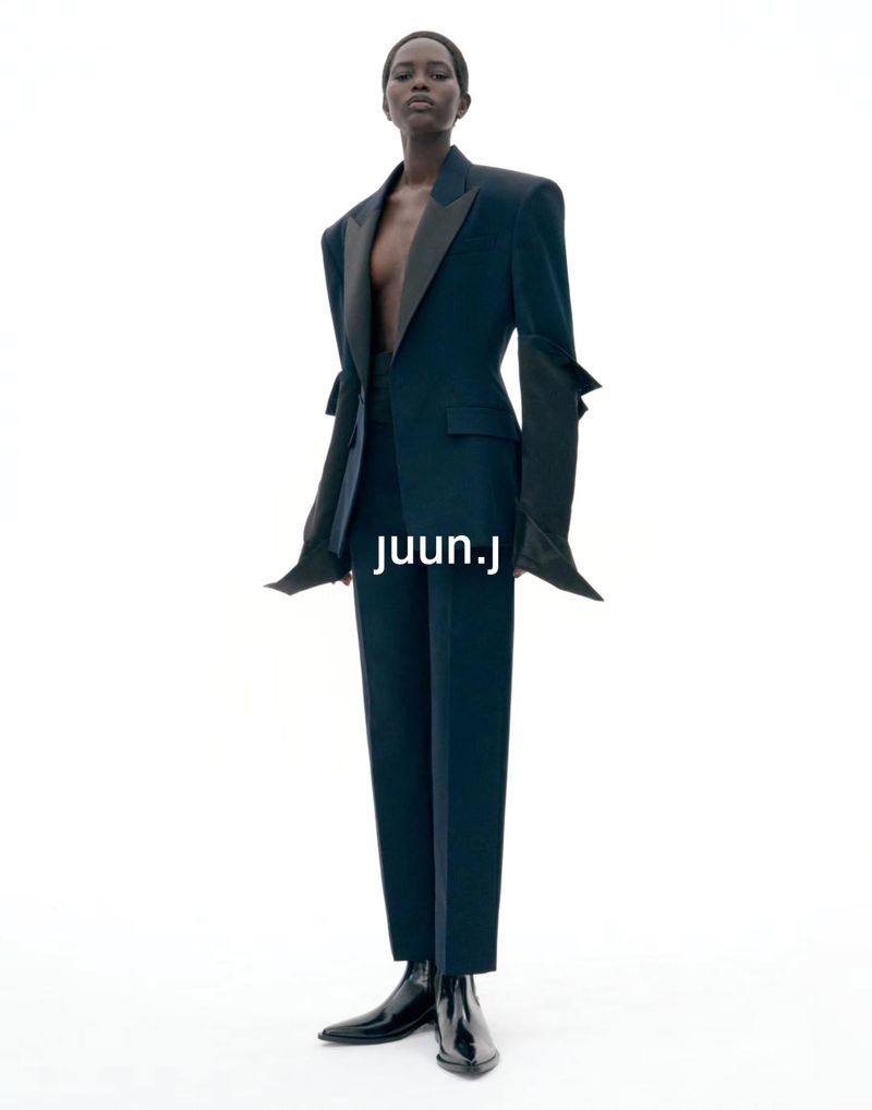 Model wearing dark structured blazer and matching trousers against plain white seamless background