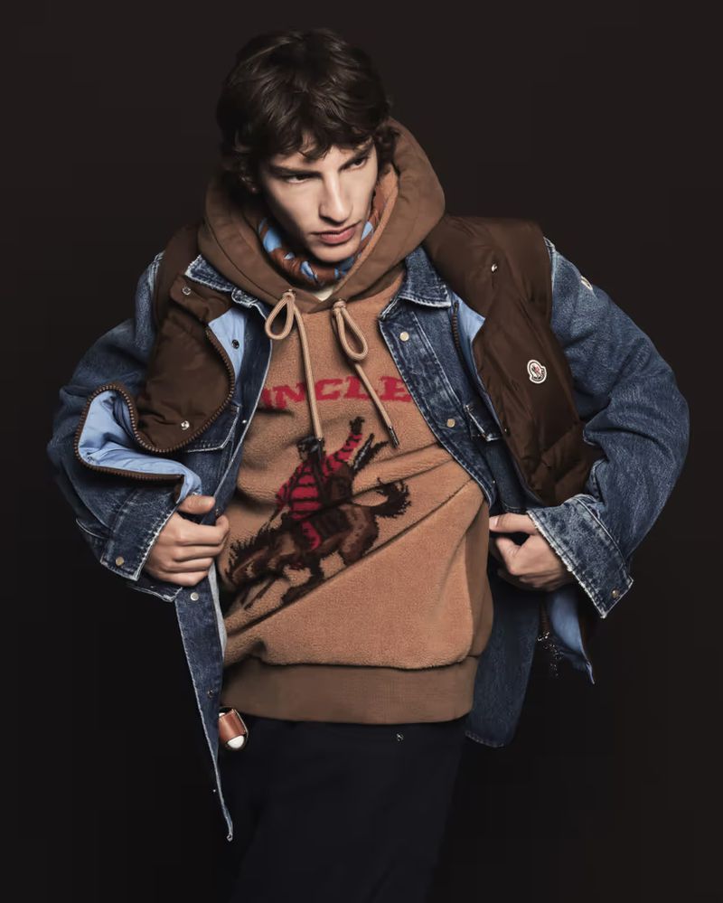 Model wearing brown graphic hoodie under denim jacket and brown vest against dark background