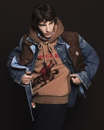 Model wearing brown graphic hoodie under denim jacket and brown vest against dark background