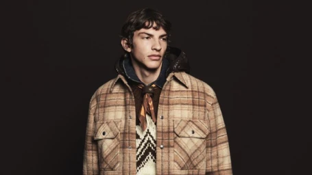 Model wearing a plaid jacket and patterned sweater against a dark background for Moncler Fall Winter 2026