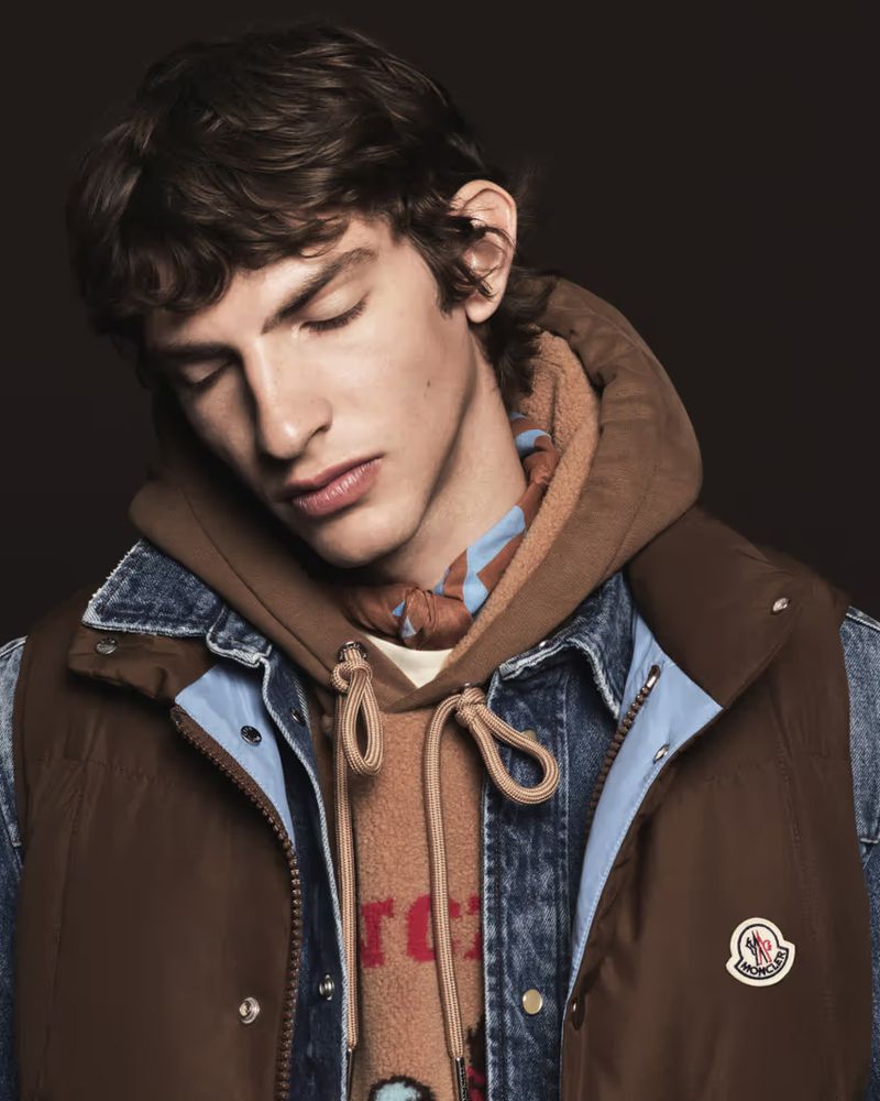 Close-up portrait of a male model wearing a brown vest over a denim jacket and hoodie with a patterned scarf against a dark background.