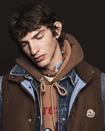 Close-up portrait of a male model wearing a brown vest over a denim jacket and hoodie with a patterned scarf against a dark background.