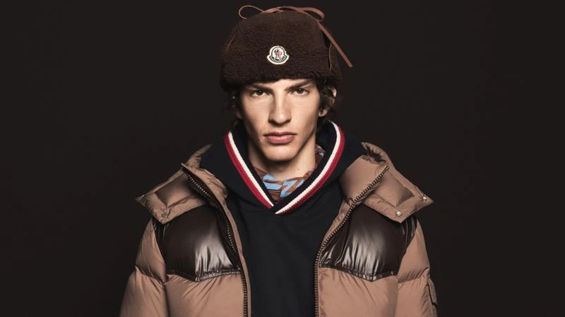 Close-up portrait of a male model wearing a brown shearling trapper hat, dark hoodie, and brown puffer jacket against a dark background.