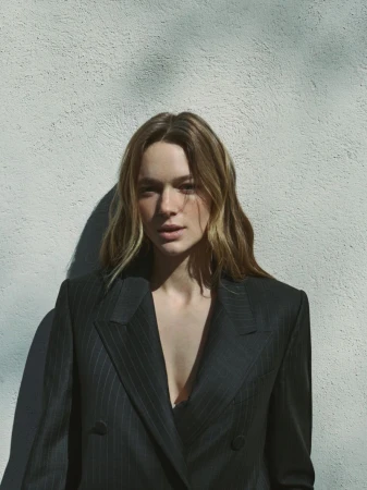 Model wearing a dark pinstripe blazer against a textured grey wall with natural shadow patterns.