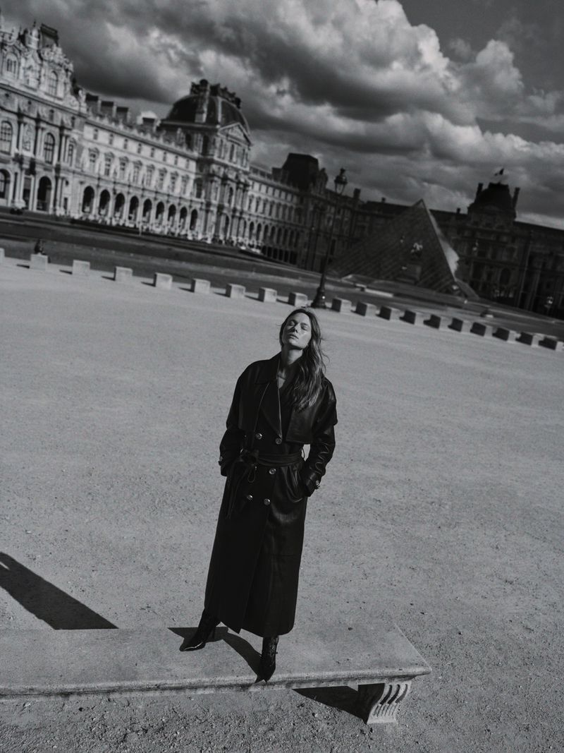 Model standing on a low stone bench in a long black coat with a historic European building facade and dramatic cloudy sky behind.