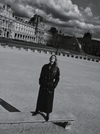 Model standing on a low stone bench in a long black coat with a historic European building facade and dramatic cloudy sky behind.