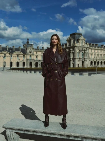 Model wearing a long burgundy leather trench coat standing on a stone bench in front of a classical building under a blue sky.