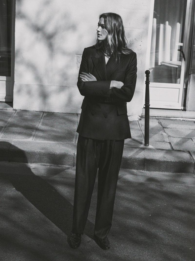 Monochrome fashion portrait of a model with arms crossed standing on a sidewalk in front of a stone building exterior.
