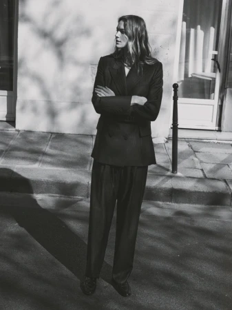 Monochrome fashion portrait of a model with arms crossed standing on a sidewalk in front of a stone building exterior.